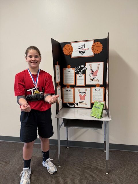 An elementary school girl in a red shirt, shows off her basketball themed engineering project.