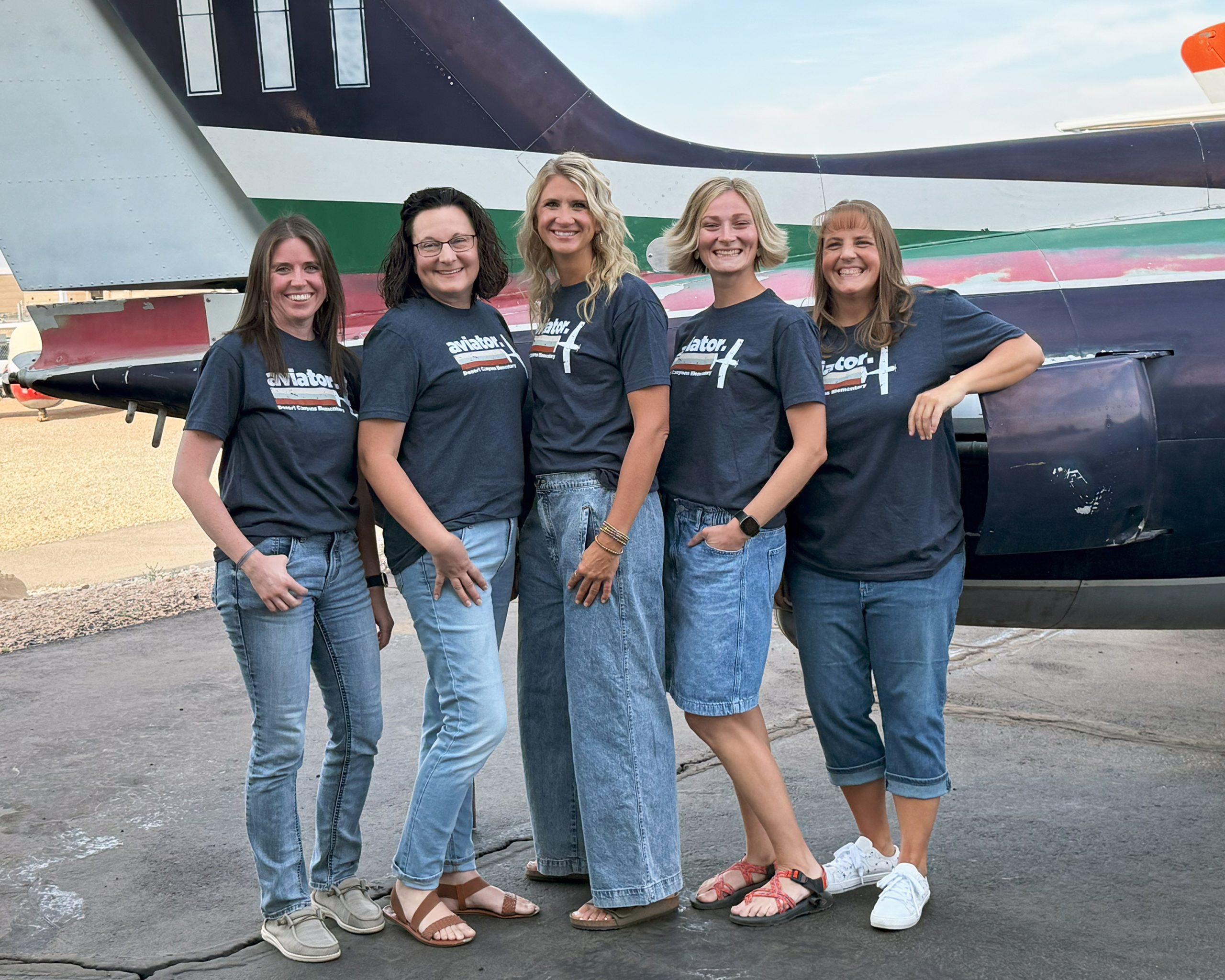 5 women pose in front of a blue, white red and green plane