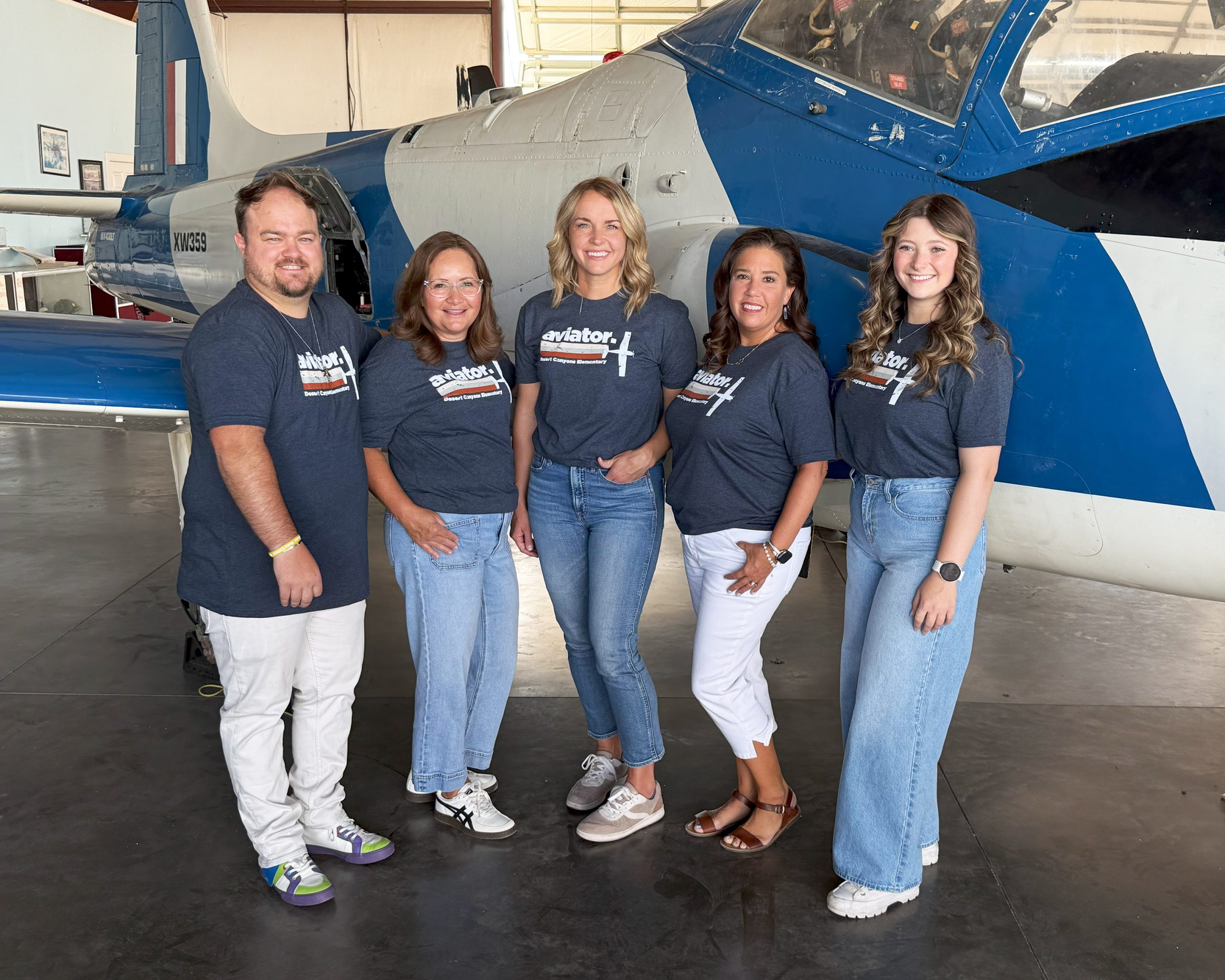 4 women and 1 man pose in front of a grey and blue airplane