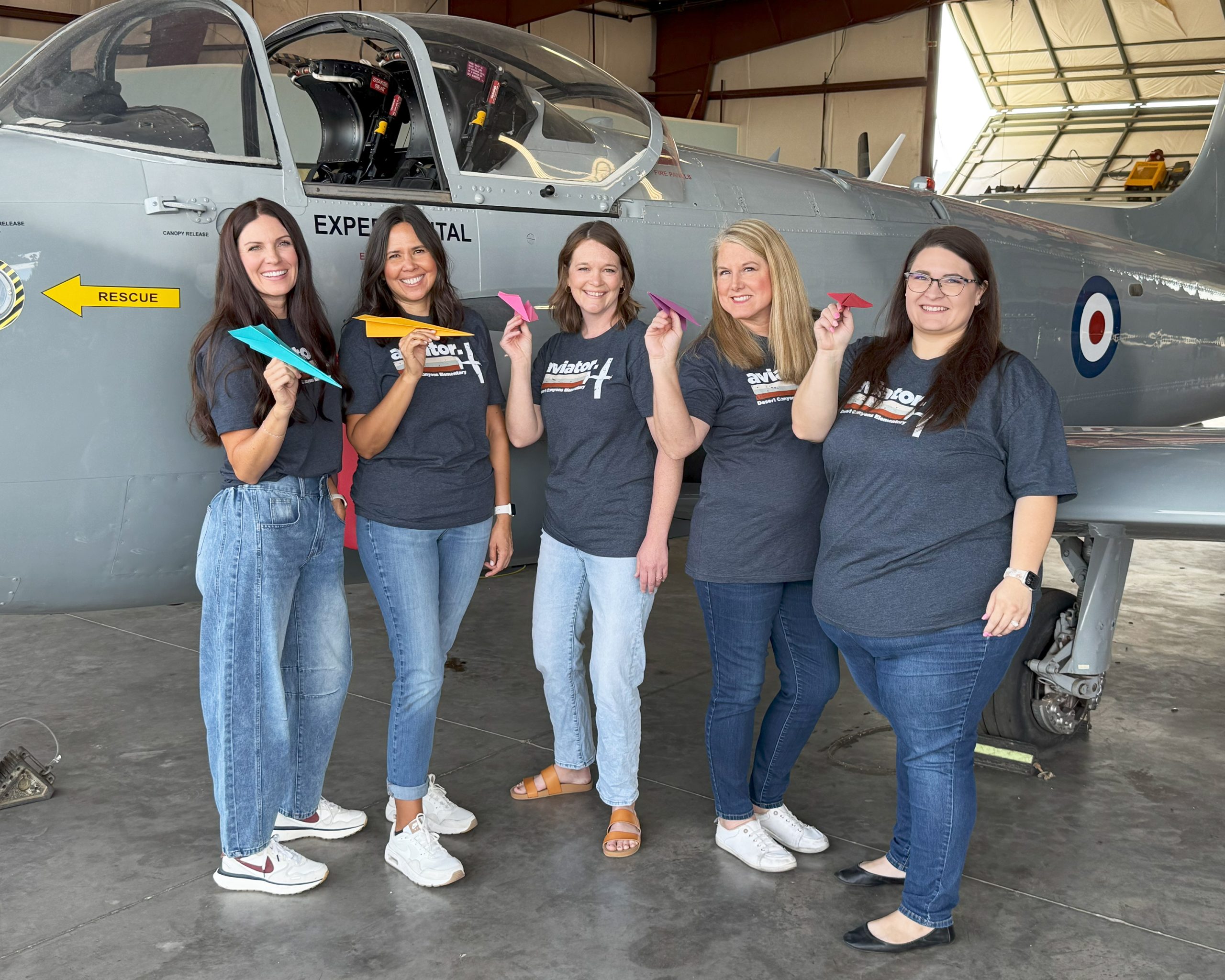 5 women pose in front of a grey airplane all holding a paper airplane