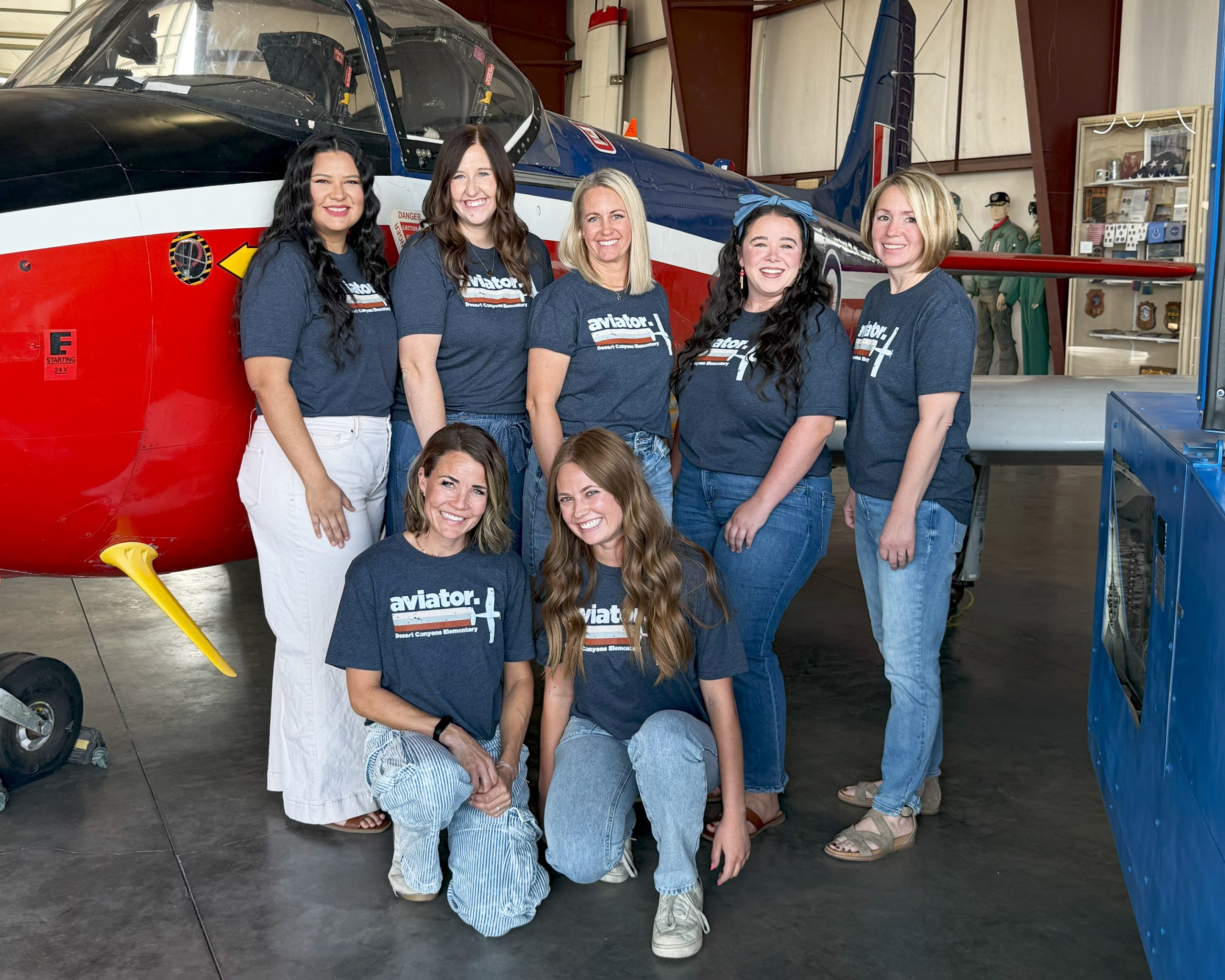 7 women pose in front of an red, white and blue plane in an airplane hangar