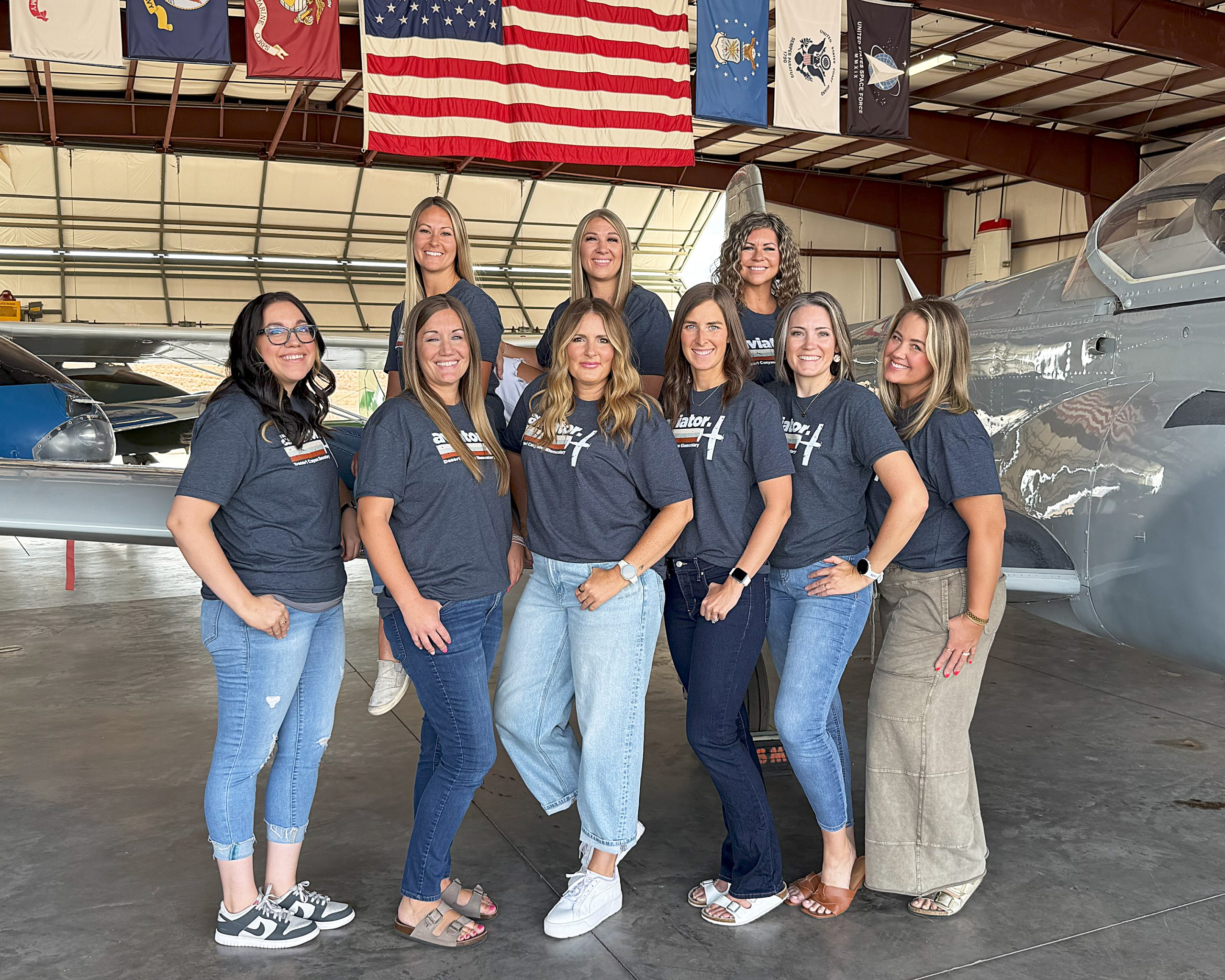 9 women pose in front of a grey airplane in an airplane hangar.
