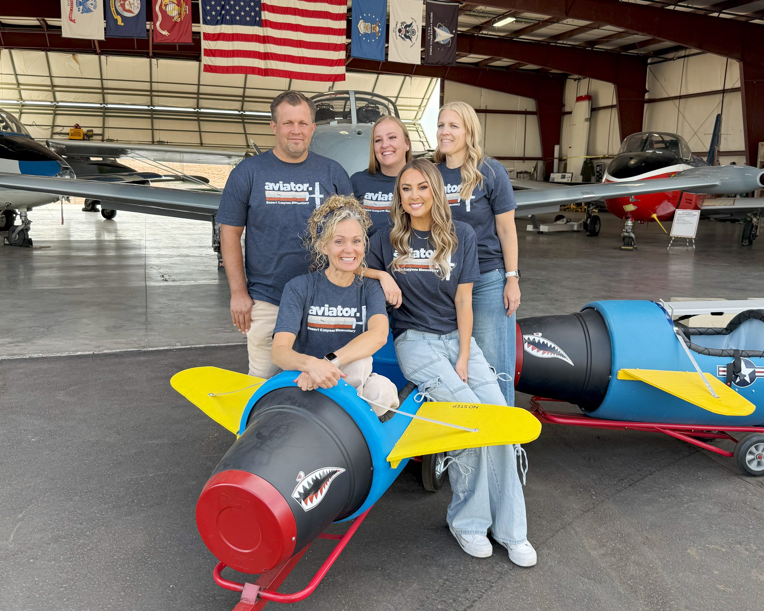 4 women and 1 man pose in front of an airplane hanger in a toy airplane