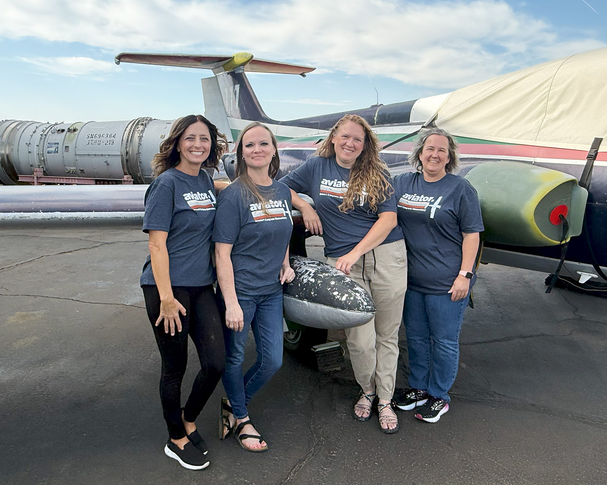 4 women pose in front of an airplane hangar with aviator shirts on
