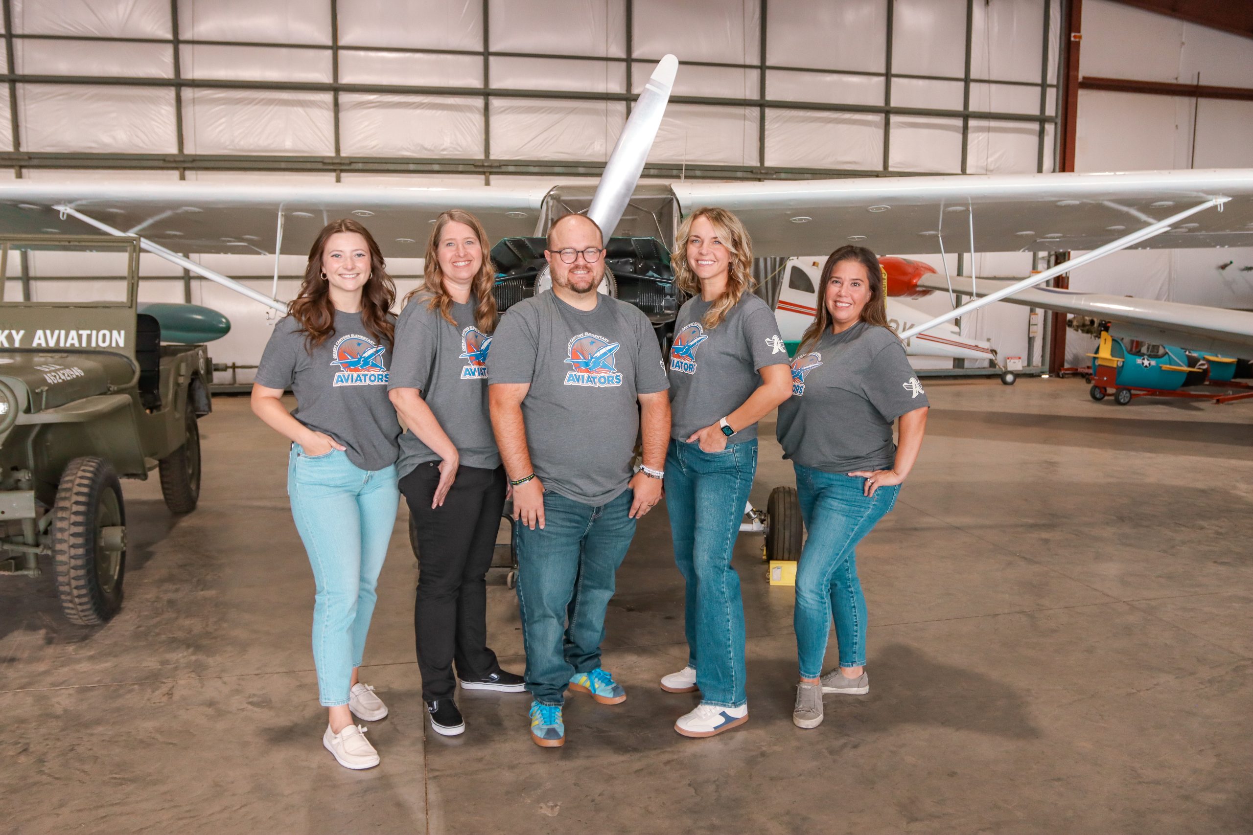 A group of 4 women and a man with matching grey school t-shirts posing next to a white airplane