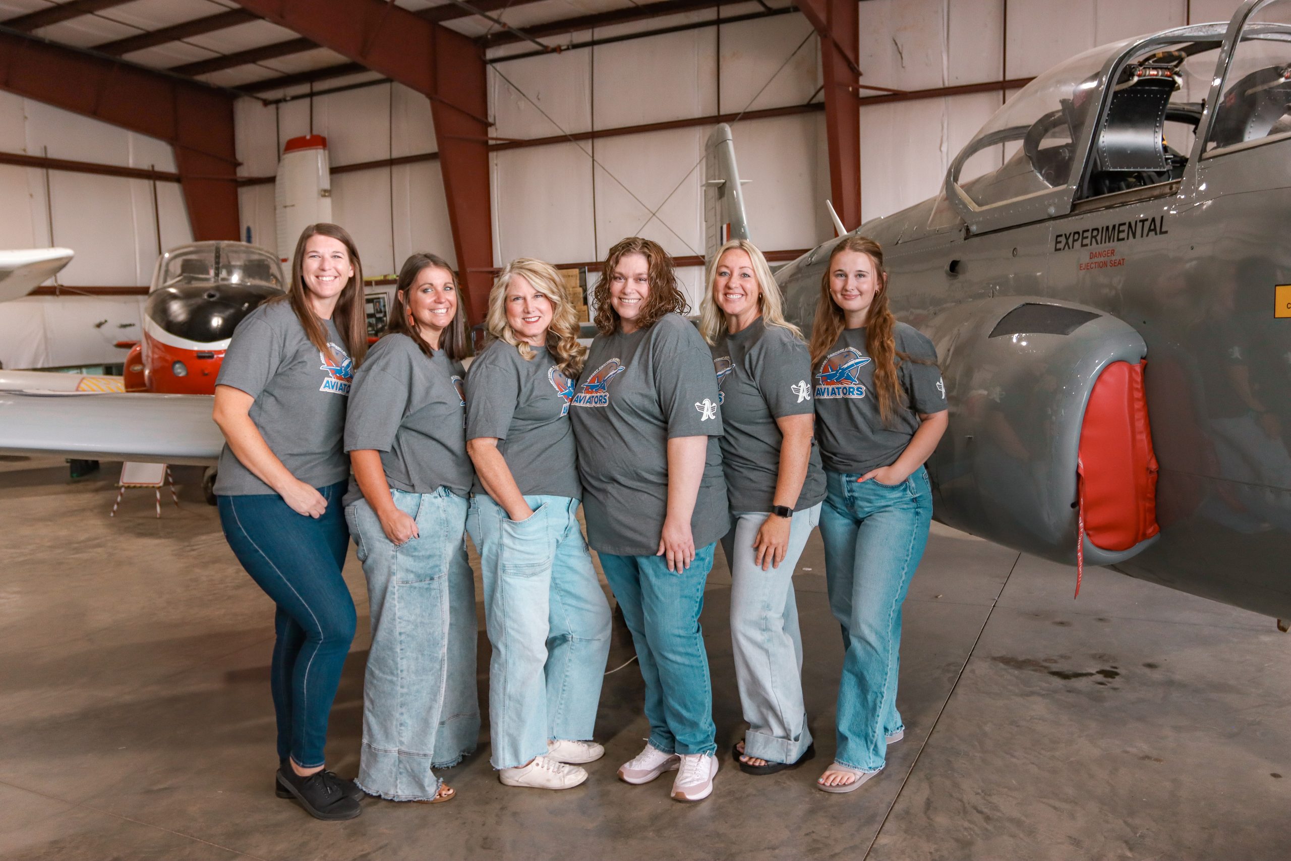 A group of 6 women with matching grey school t-shirts posing next to a grey airplane