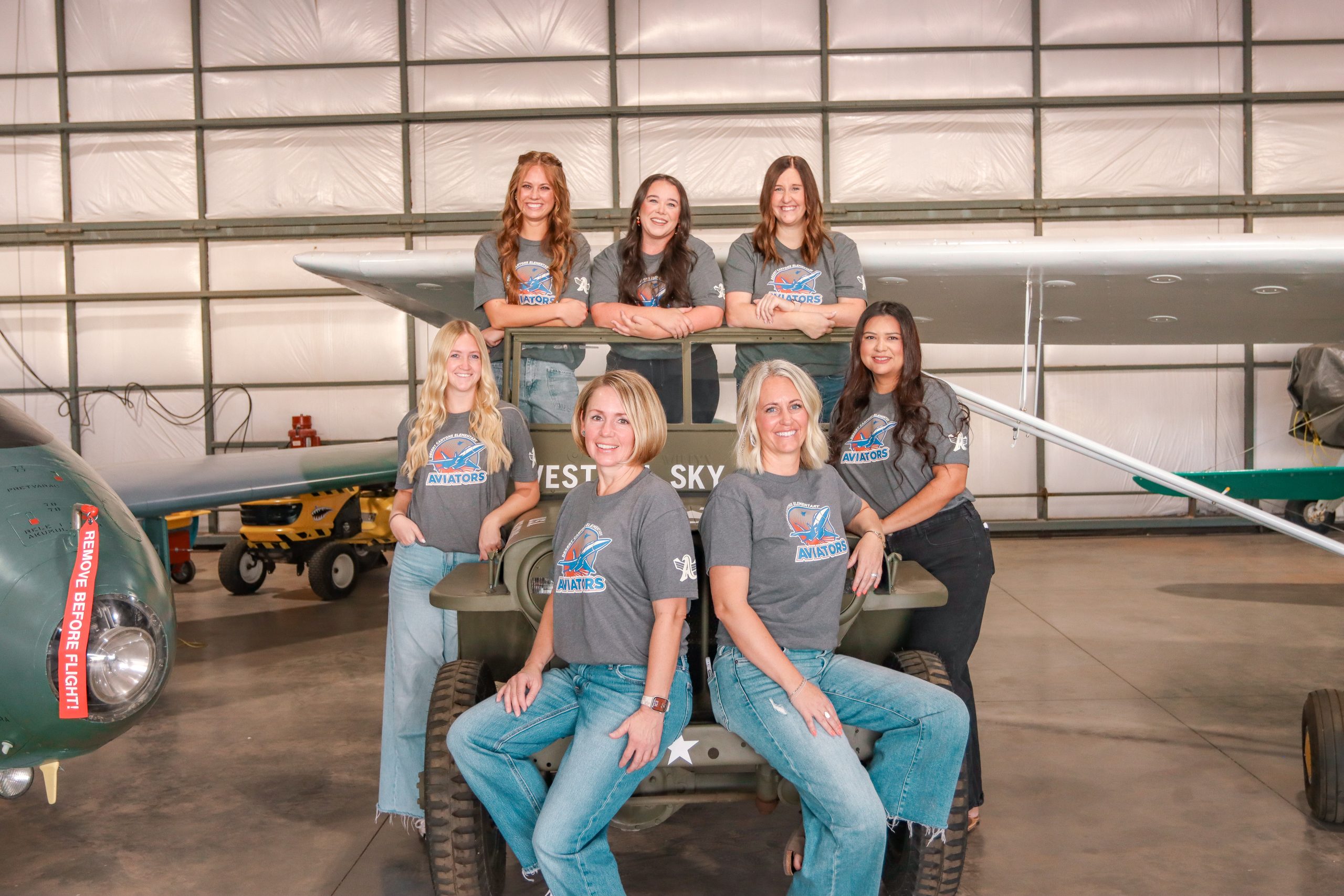 A group of 7 women with matching grey school t-shirts posing next to and on top of a green military jeep with airplanes around them
