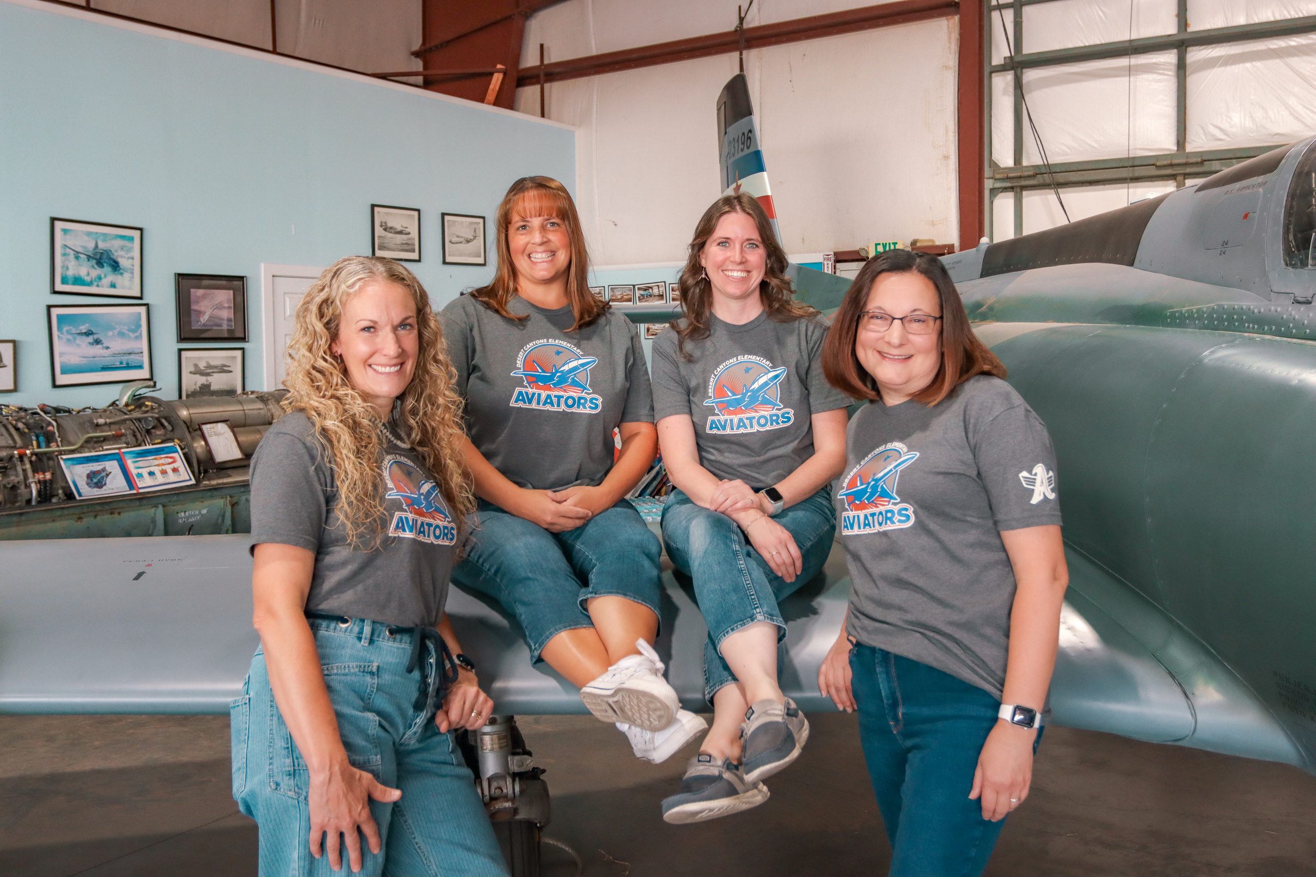 A group of 4 women with matching grey school t-shirts posing next to and on top of a grey airplane in a hangar