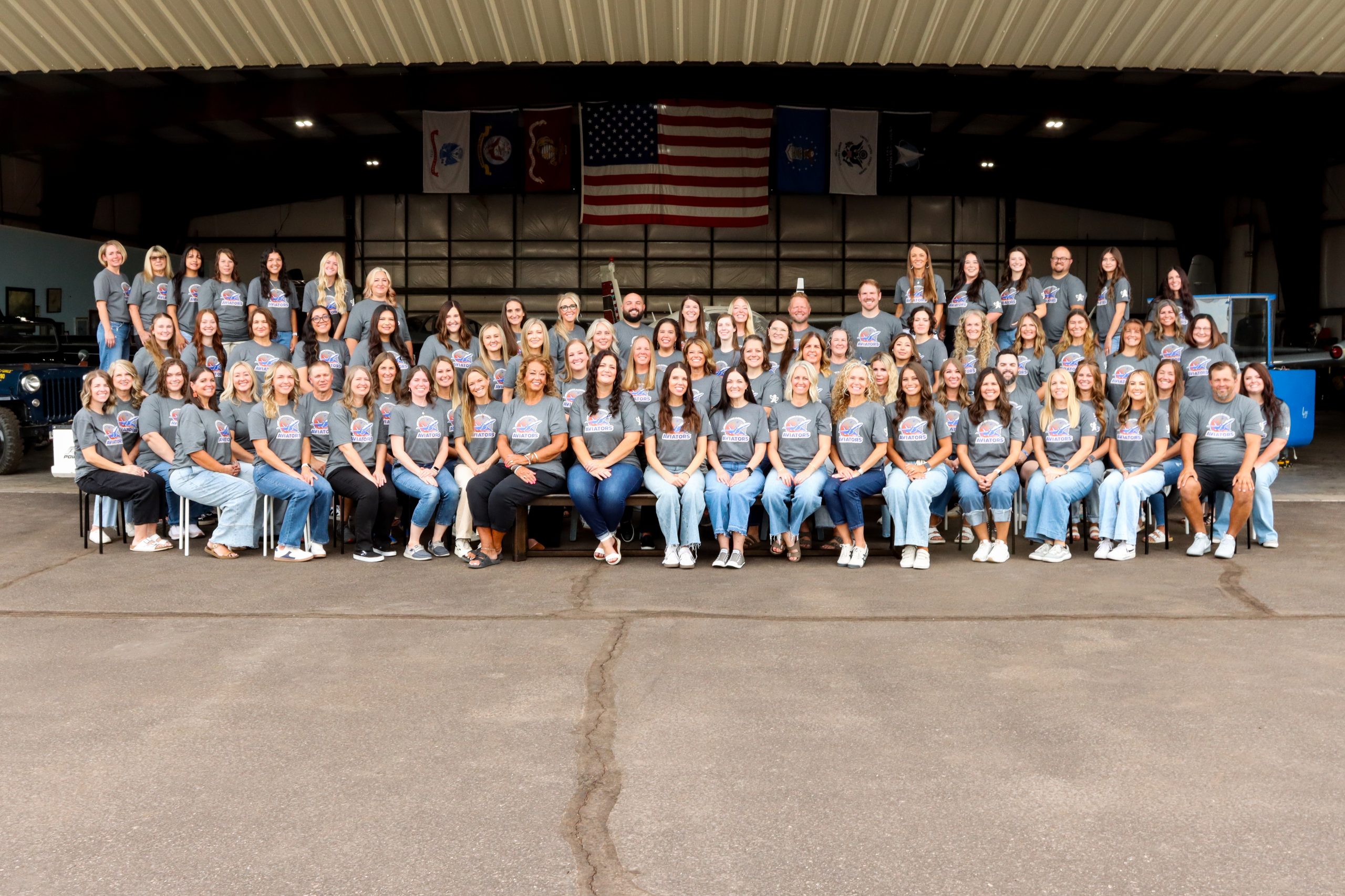 A very large group of people with matching grey school t-shirts pose in front of an airplane hangar