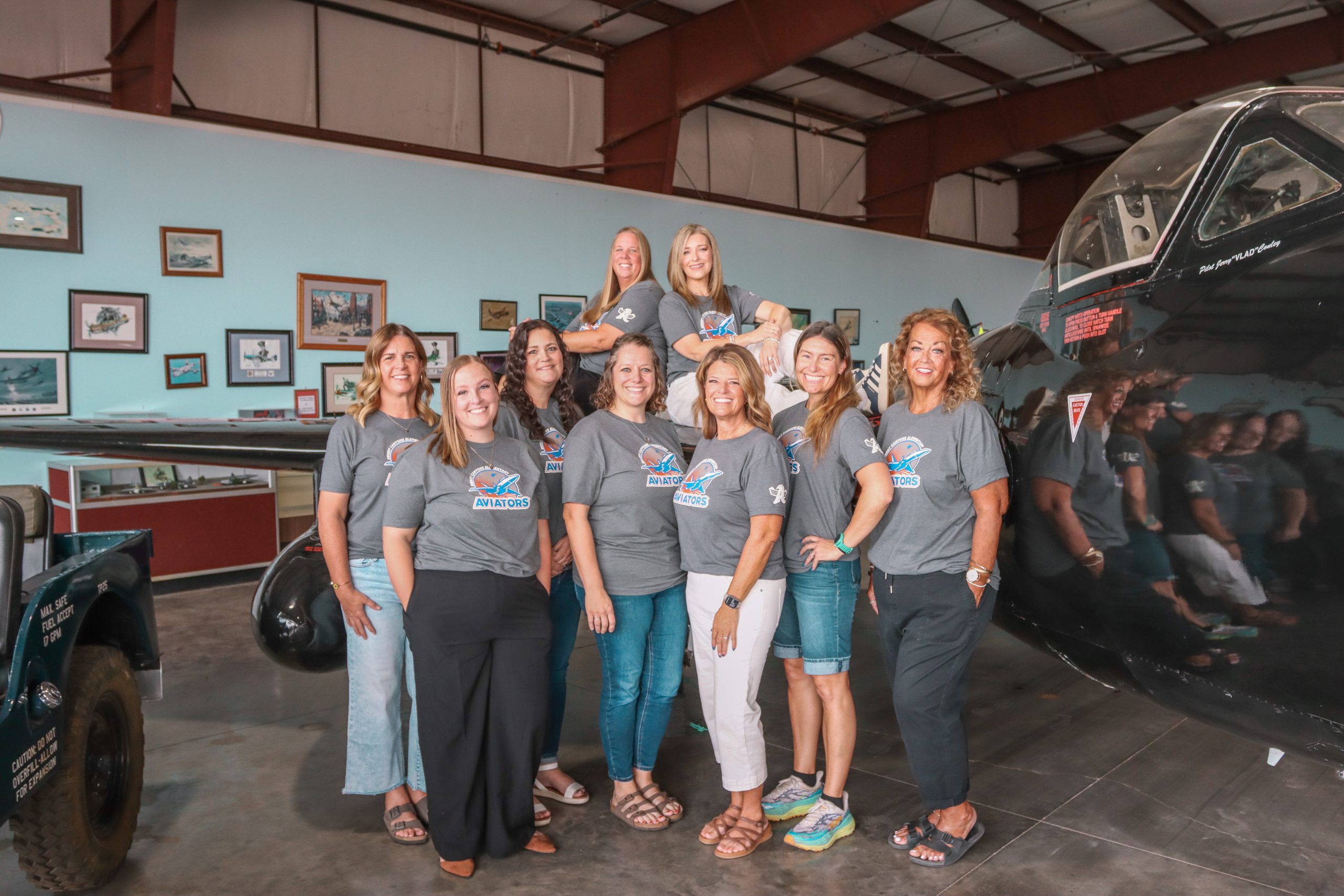 a group of 12 women, matching grey school t-shirts pose near and on top of a black airplane