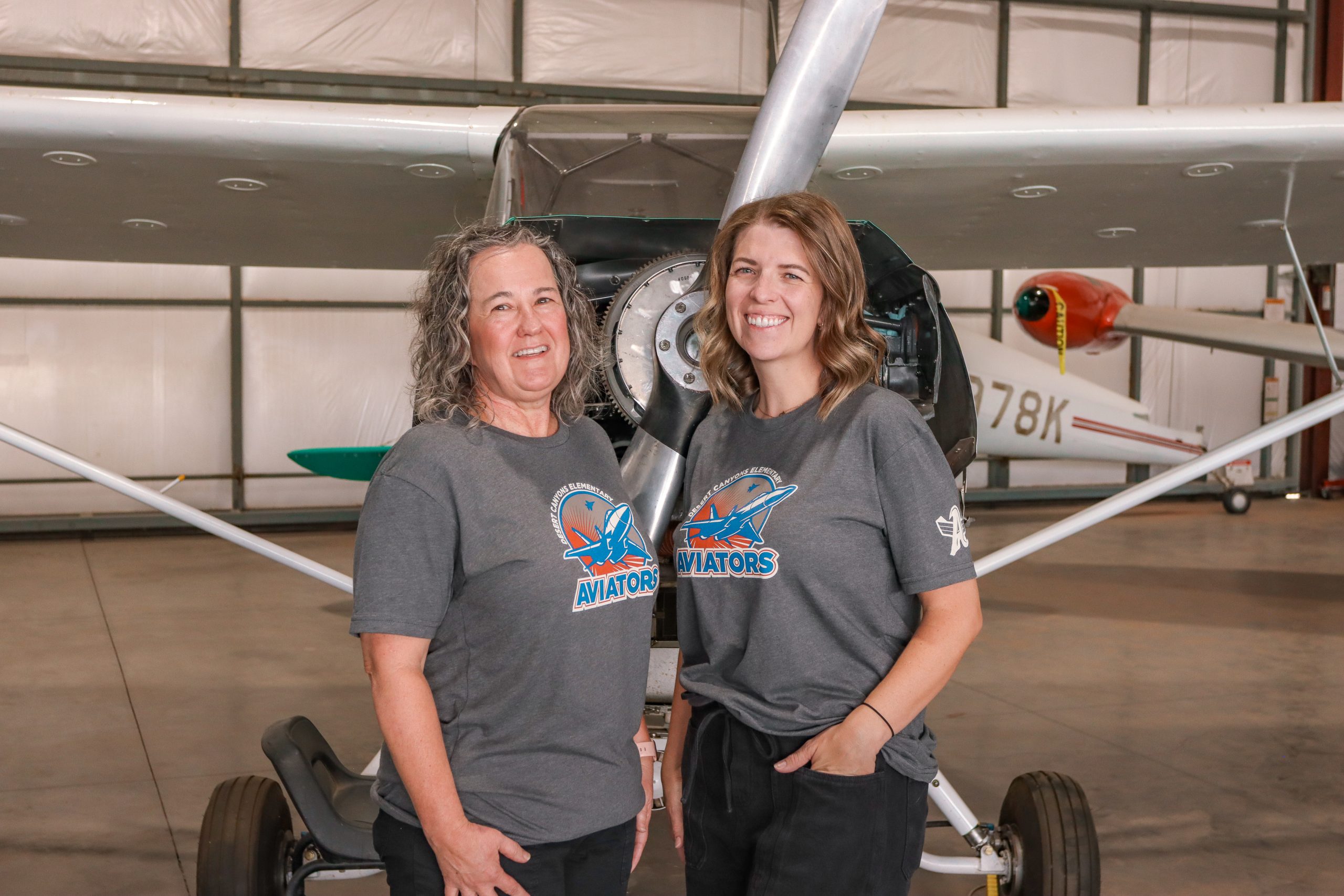 two women in matching grey school t-shirts pose in front of an airplane propellor
