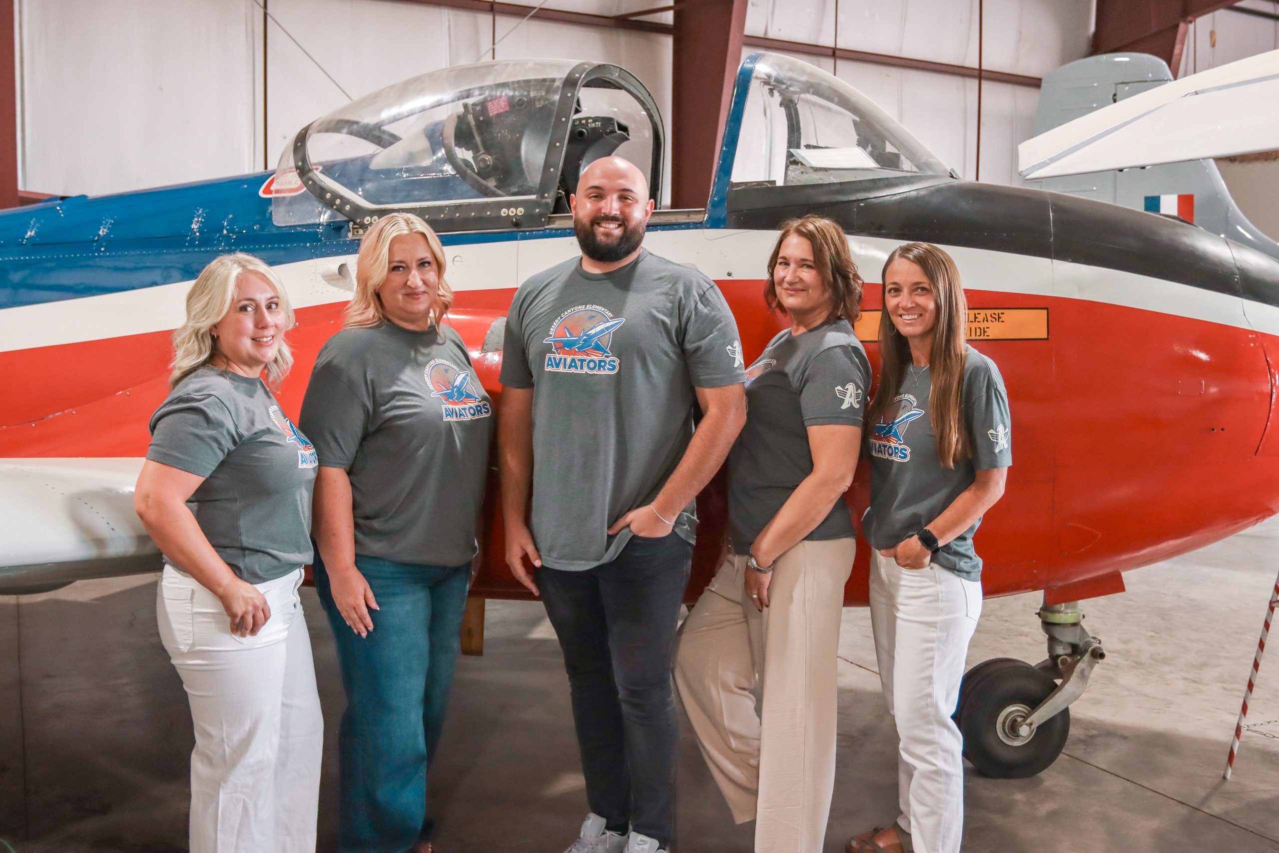 A group of 4 women and a man with matching grey school t-shirts, pose in front of a red, white and blue airplane