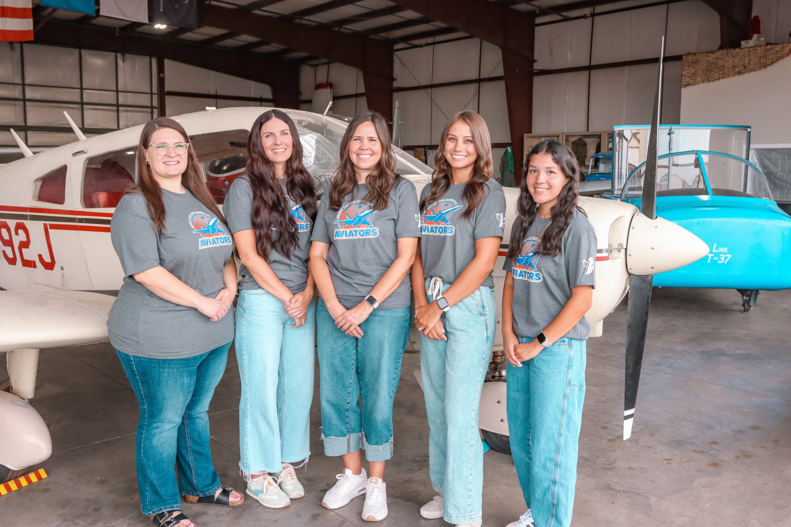 A group of 5 women in matching school t-shirts pose in front of a white airplane