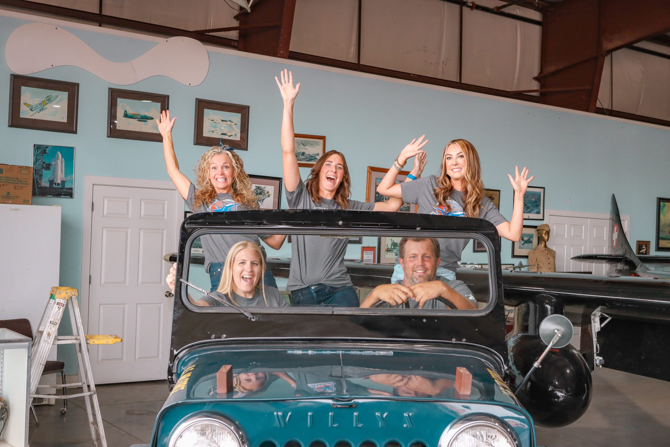 A group of 4 women and a man with matching grey school t-shirts posing in a military jeep. The 3 women in the back seat have their arms raised in the air