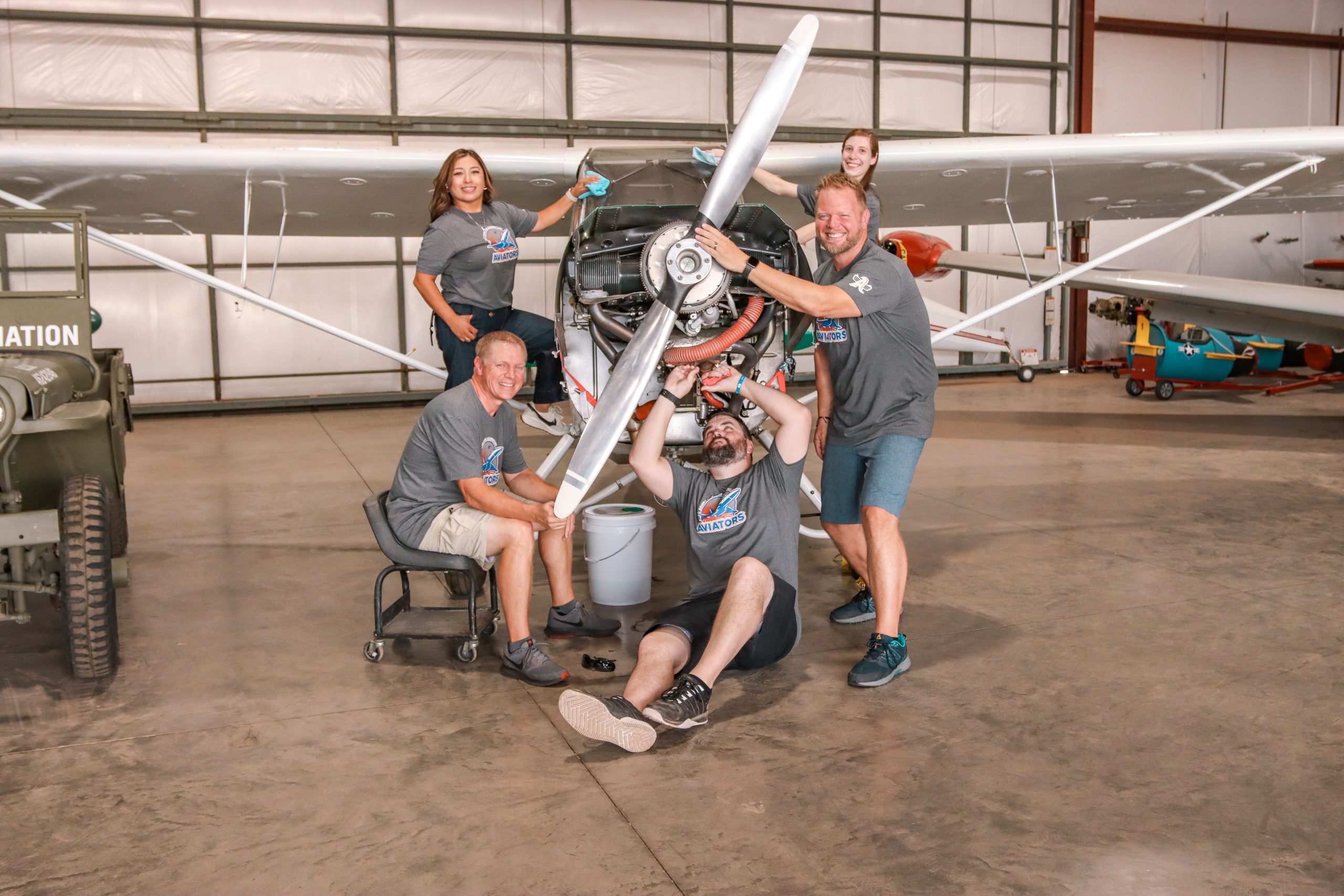 a group of 3 men and 2 women with matching grey school t-shirts pretending to fix an airplane in a hangar
