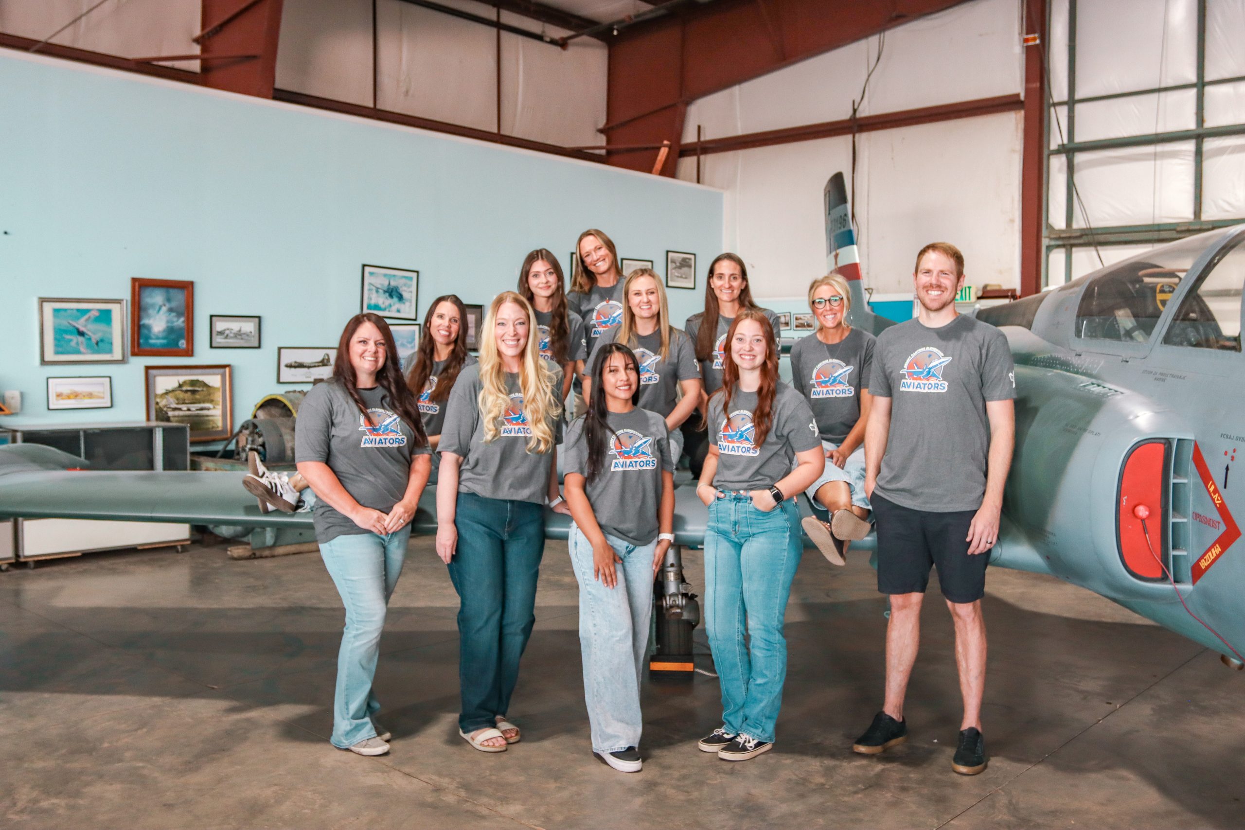 A group of 10 women and a man in matching school t-shirts posing near a grey airplane